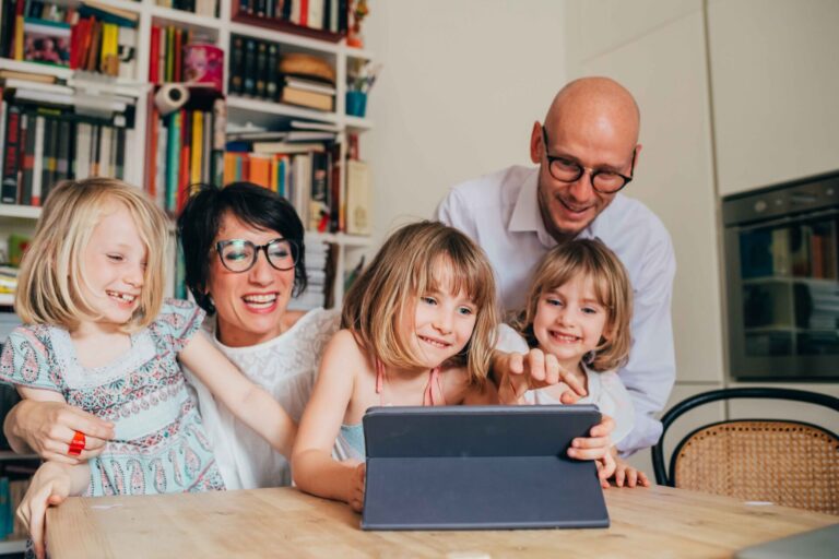 Three little sisters indoor at home using tablet sitting table supervised by parents