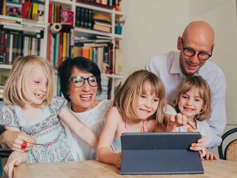 three girls and their parents look at schoolwork on a tablet together.