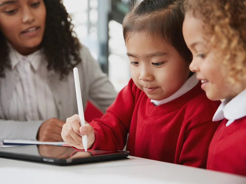 a student shows her work on a tablet while a classmate and teacher watch