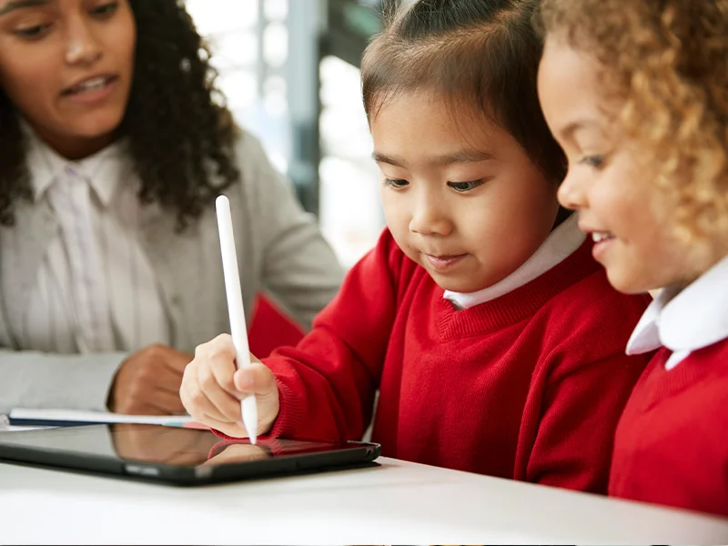 a student shows her work on a tablet while a classmate and teacher watch
