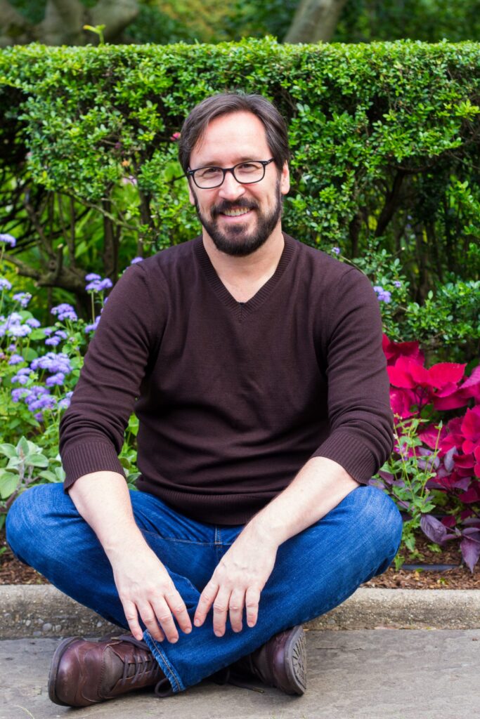 Author Wiley Blevins, a man with a beard and glasses sitting in a garden wearing a sweater and jeans