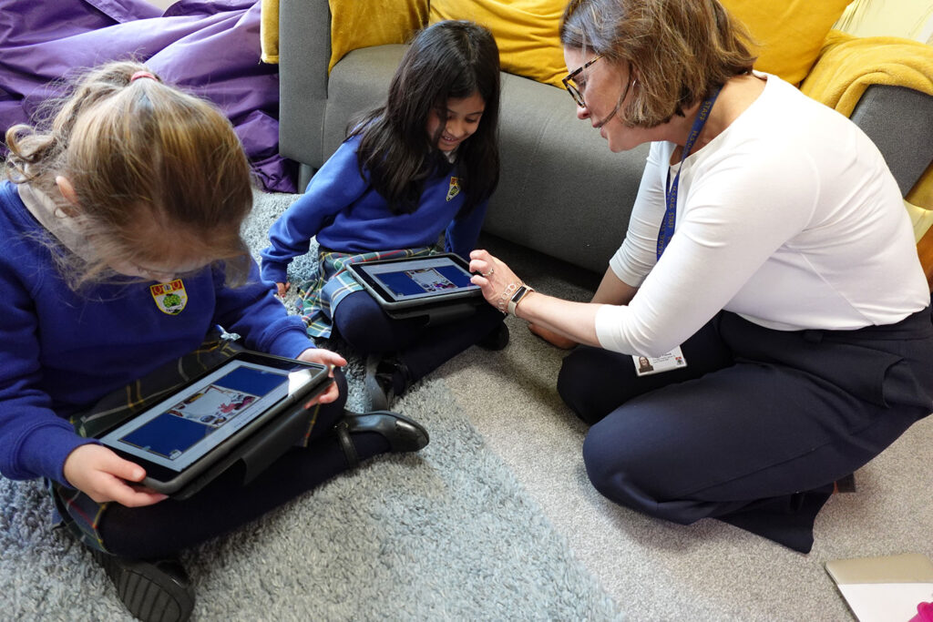 A female teacher wearing glasses kneels on a rug next to two young pupils in blue uniforms who are looking at educational apps on tablets.