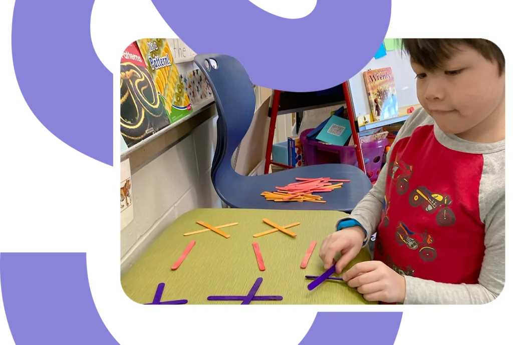 Young child in a classroom arranging orange, pink, and purple craft sticks into repeating patterns on a desk.